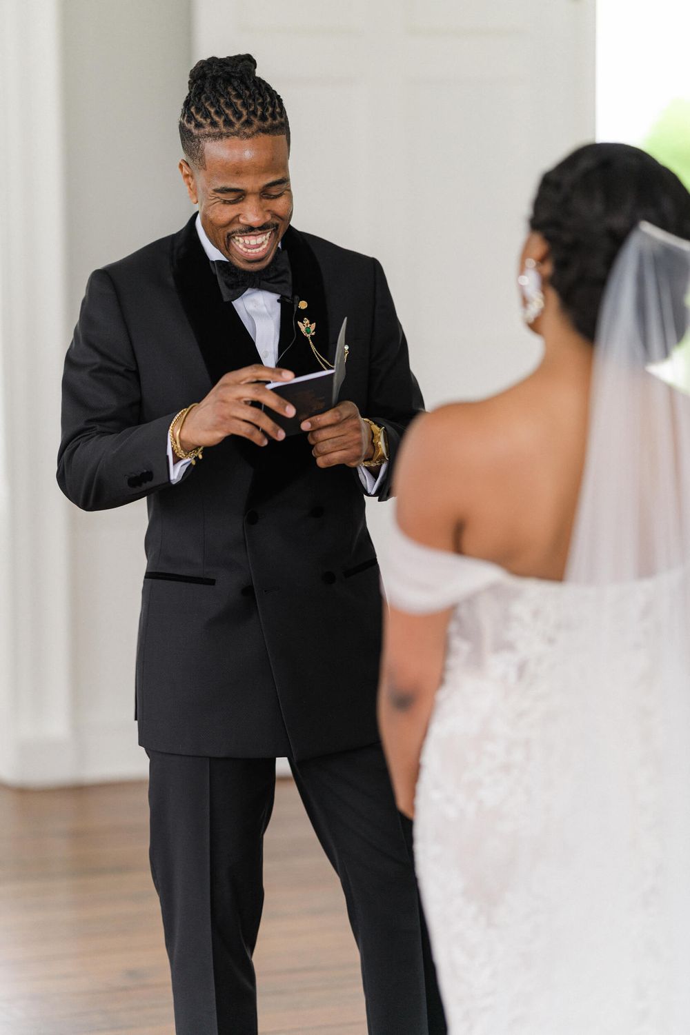 Groom reading his wedding vows with joy while bride watches in an elegant white room.