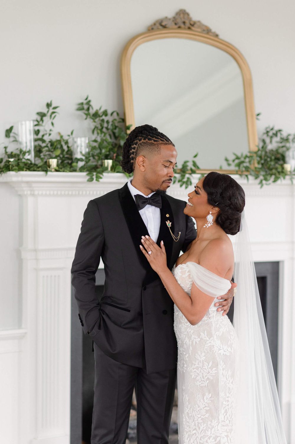 Couple shares an intimate moment in front of a decorative fireplace with gold mirror and greenery.