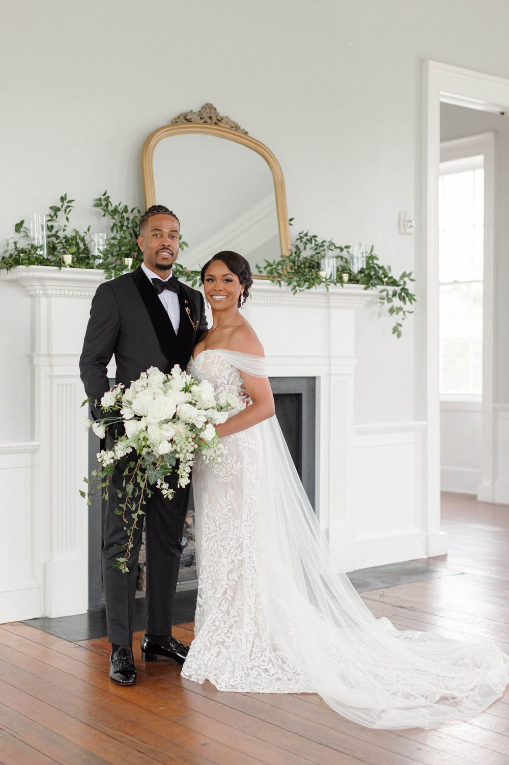 Wedding portrait of couple standing by white fireplace with ornate mirror and cascading white bouquet.