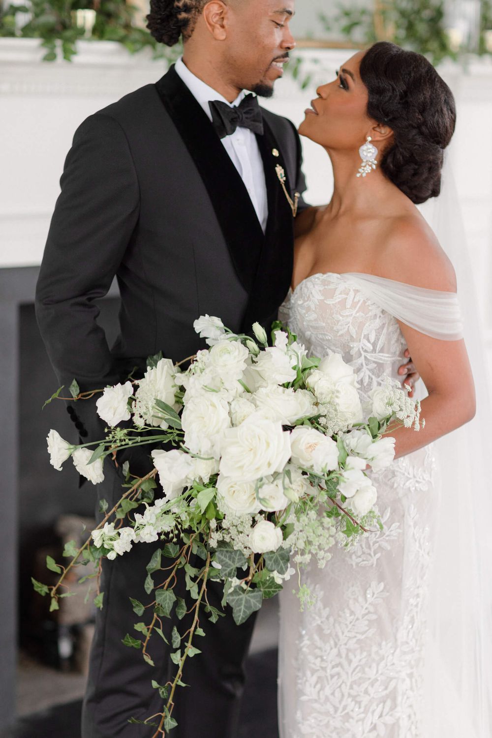 Romantic wedding portrait with bride holding a cascading white bouquet against white fireplace.