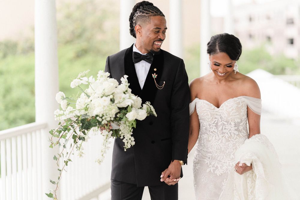 Elegant wedding couple stands on a white porch with a beautiful cascading white floral bouquet.