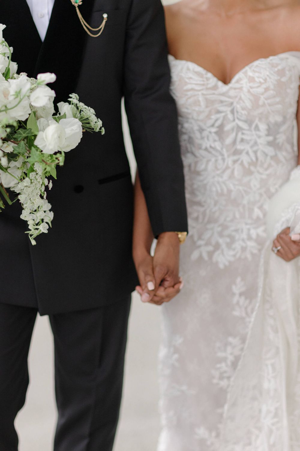Close-up of bride in lace gown and groom in tuxedo holding hands with white floral bouquet.