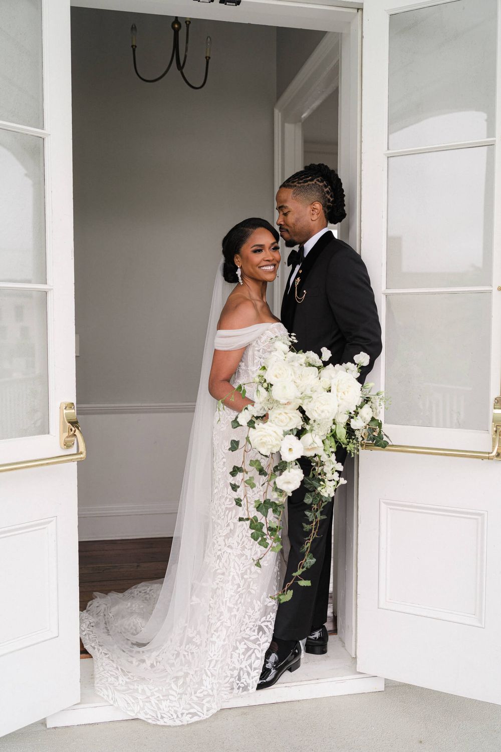 Wedding couple poses in white doorframe with cascading white floral bouquet and lace gown.