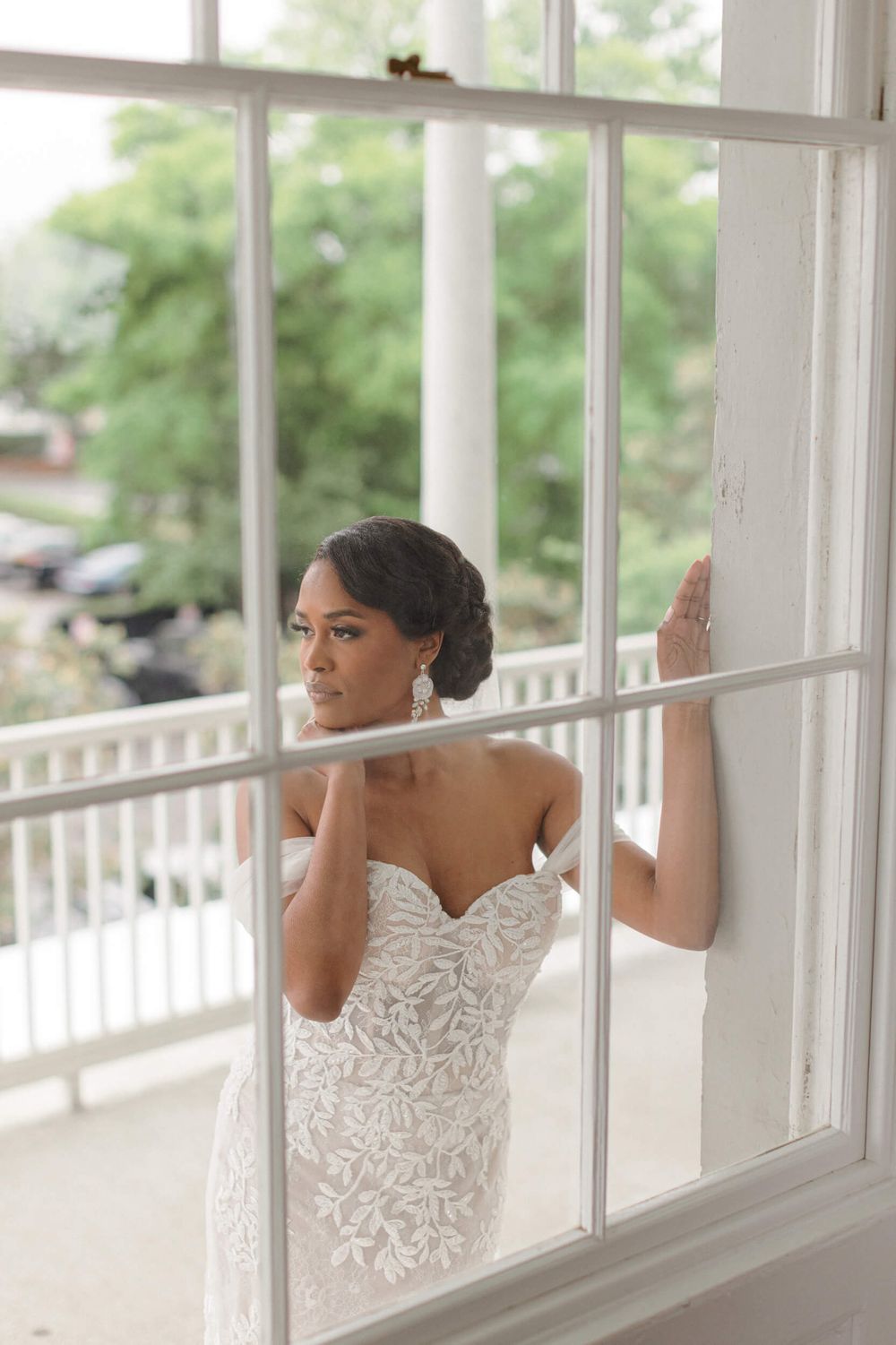 Bride in elegant lace gown gazes through white-framed windows on a bright porch.
