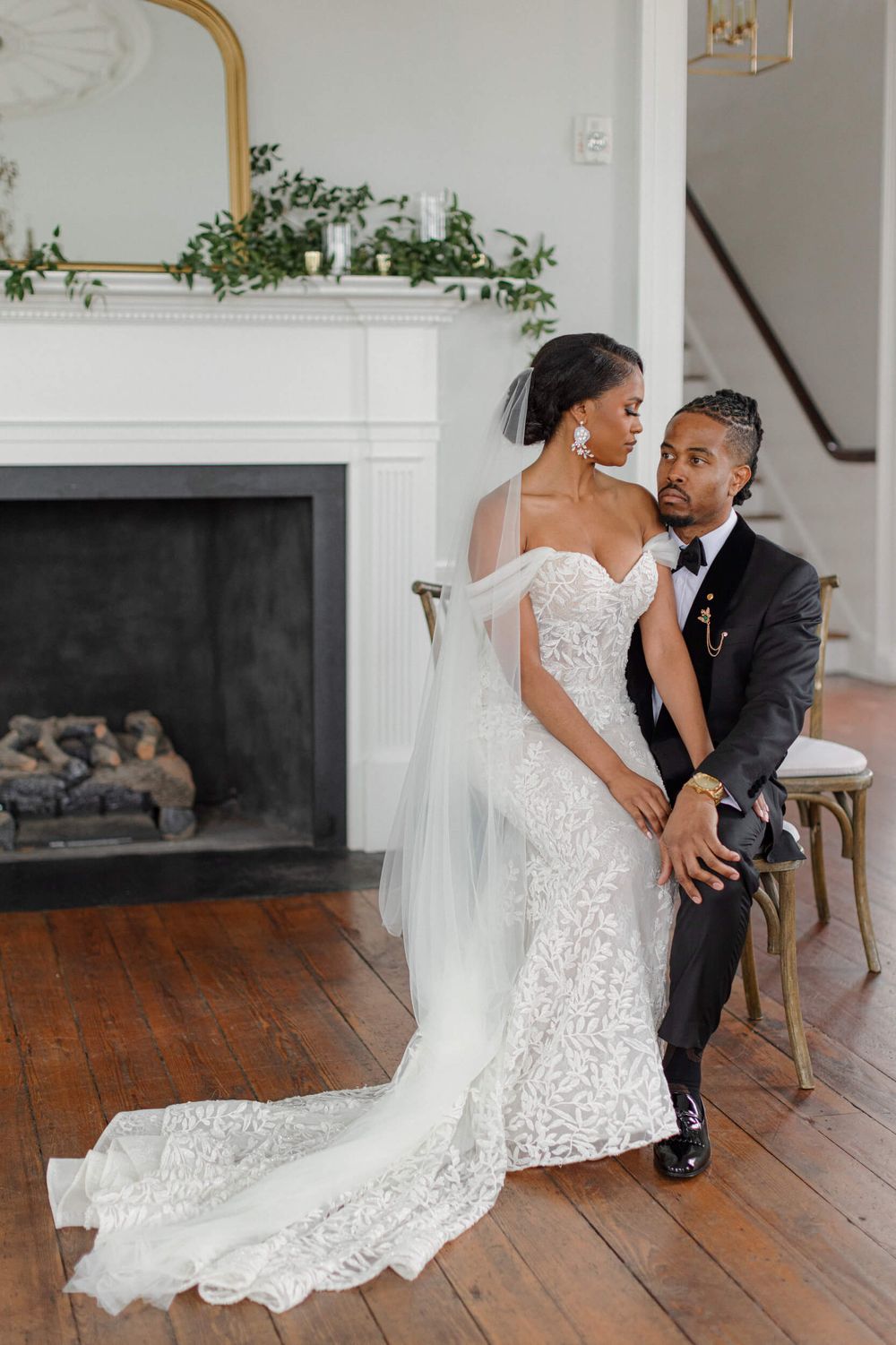 Wedding couple poses by white fireplace with greenery and elegant decor.