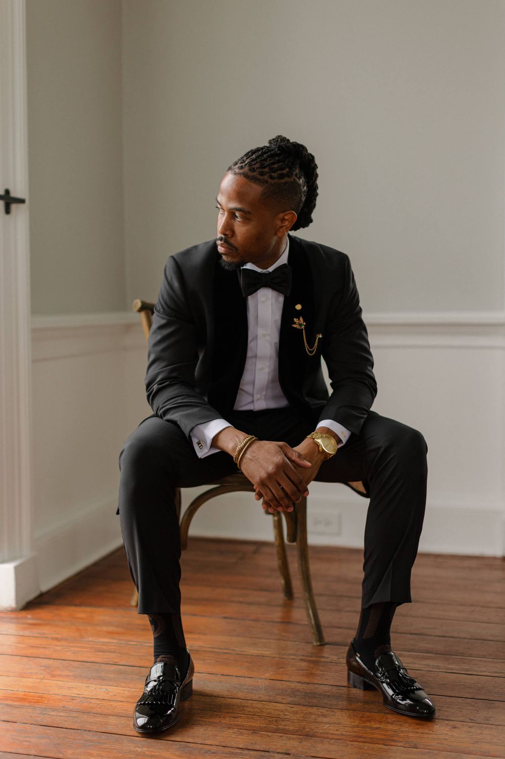 Groom in classic black tuxedo sits contemplatively on wooden chair.