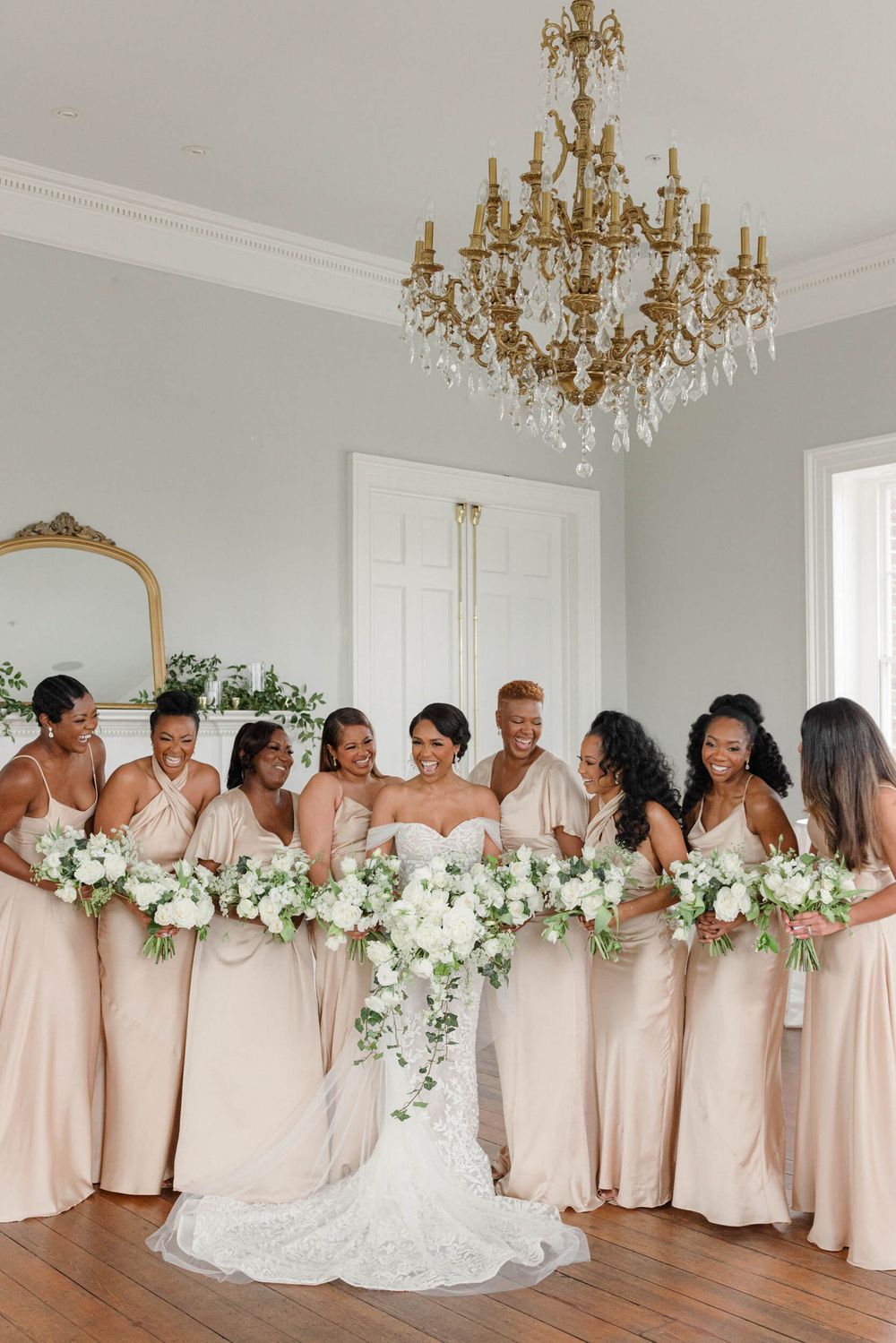 Bridal party in blush dresses poses under crystal chandelier with white floral bouquets.