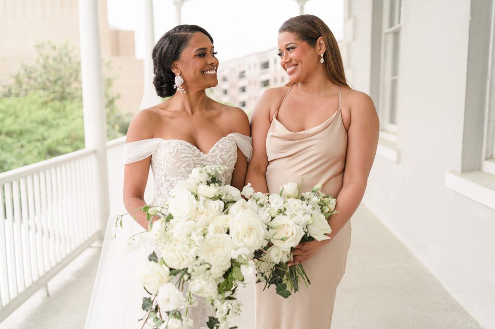 Two people in wedding attire share a joyful moment on a white porch with floral bouquets.