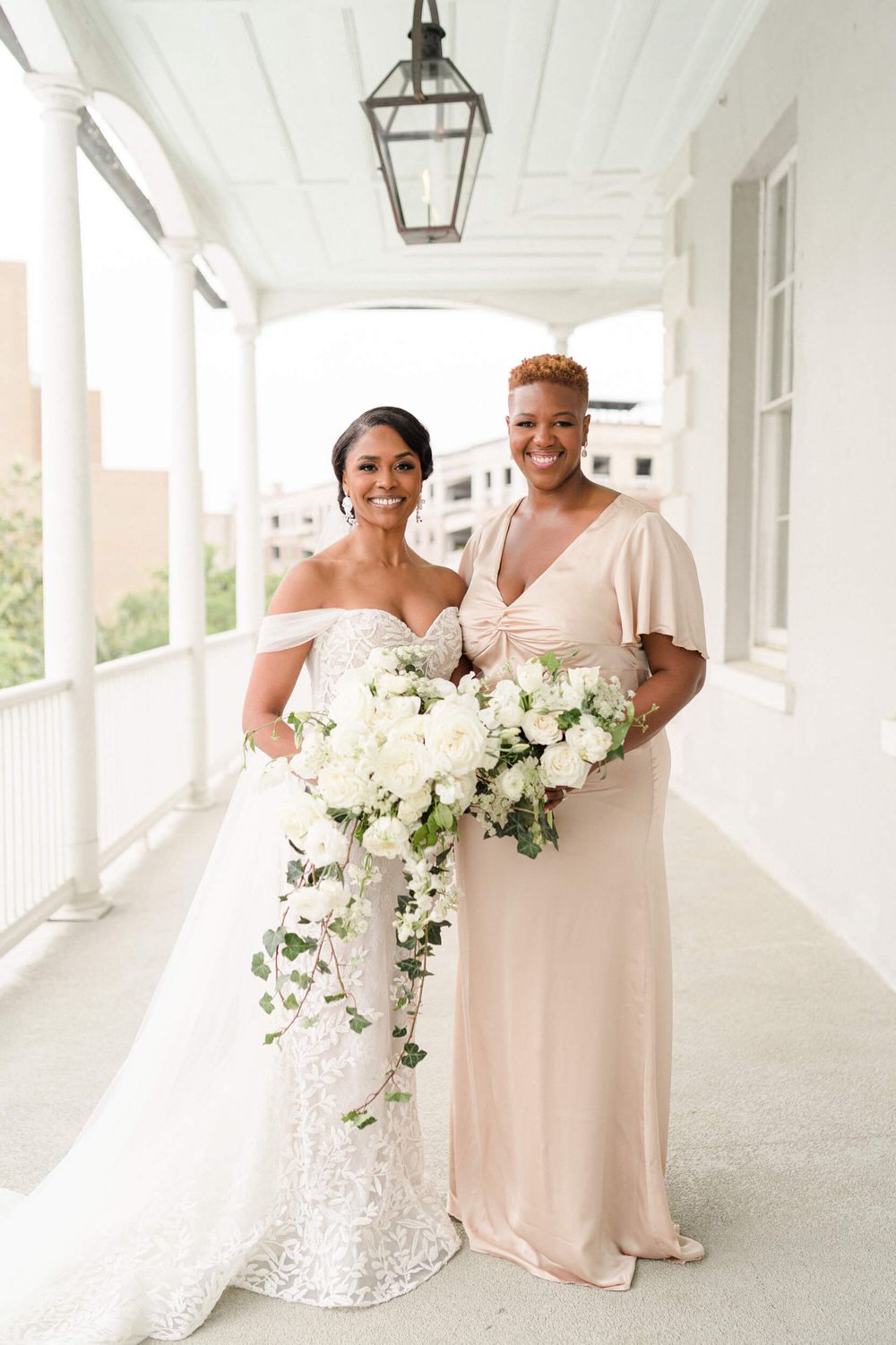 Two people in formal attire pose with white bouquets on an elegant white columned porch.