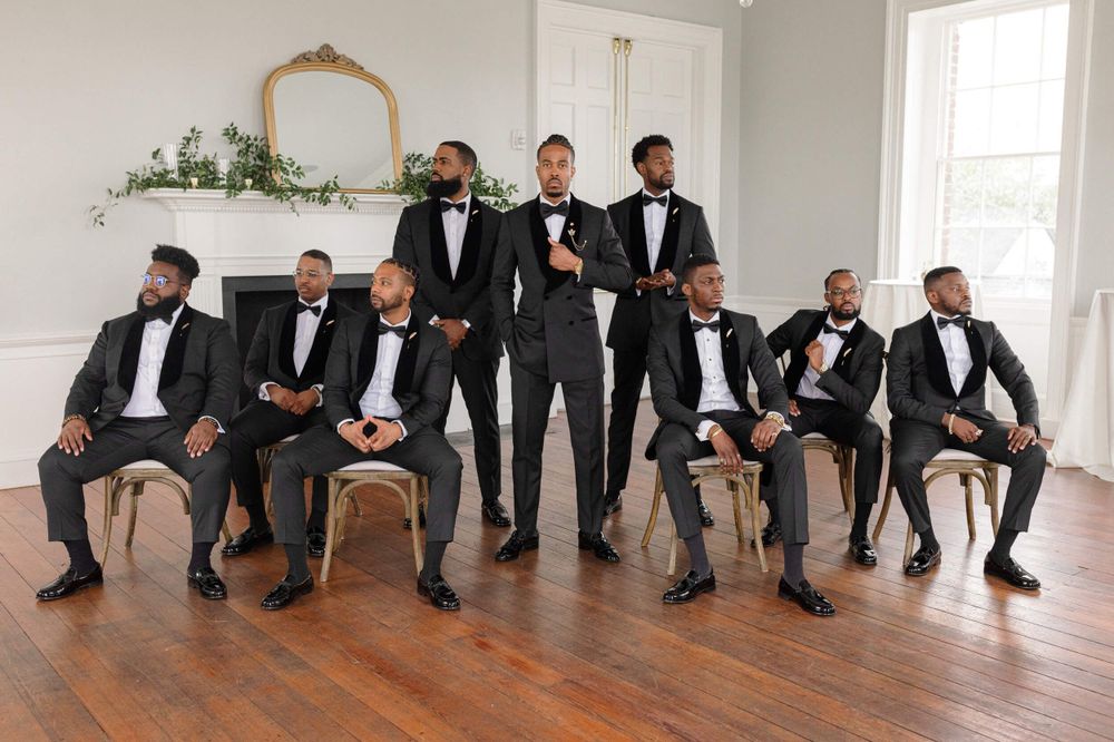 Groomsmen in black tuxedos pose for formal wedding photo in bright white room.