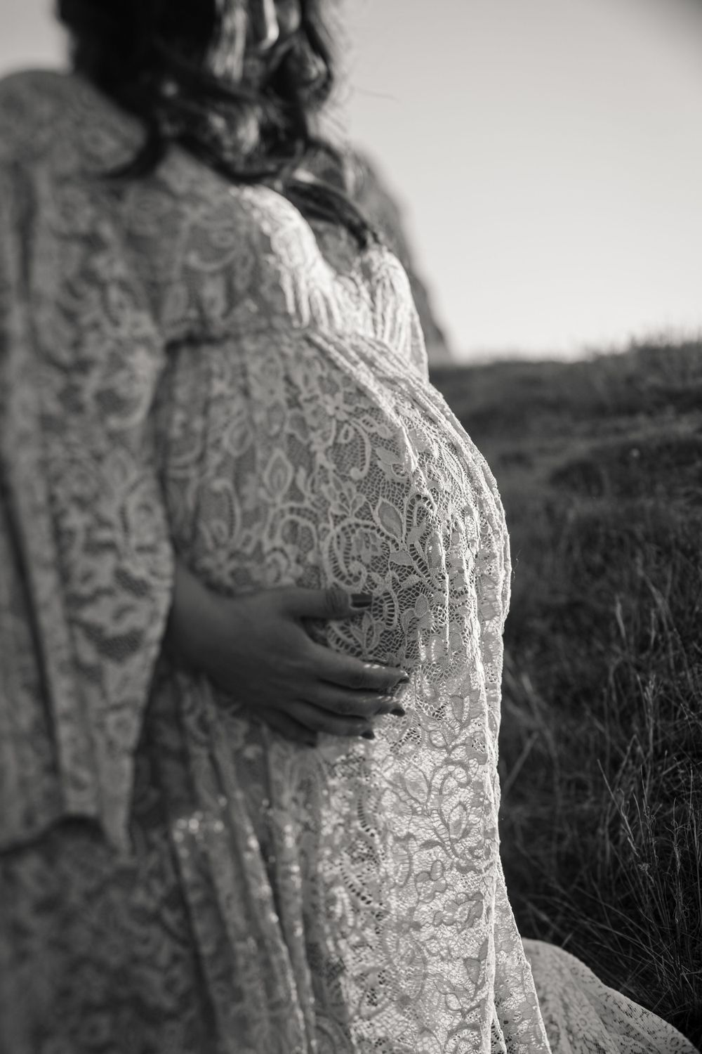 Black and white portrait highlighting delicate lace detail of maternity dress in natural setting.