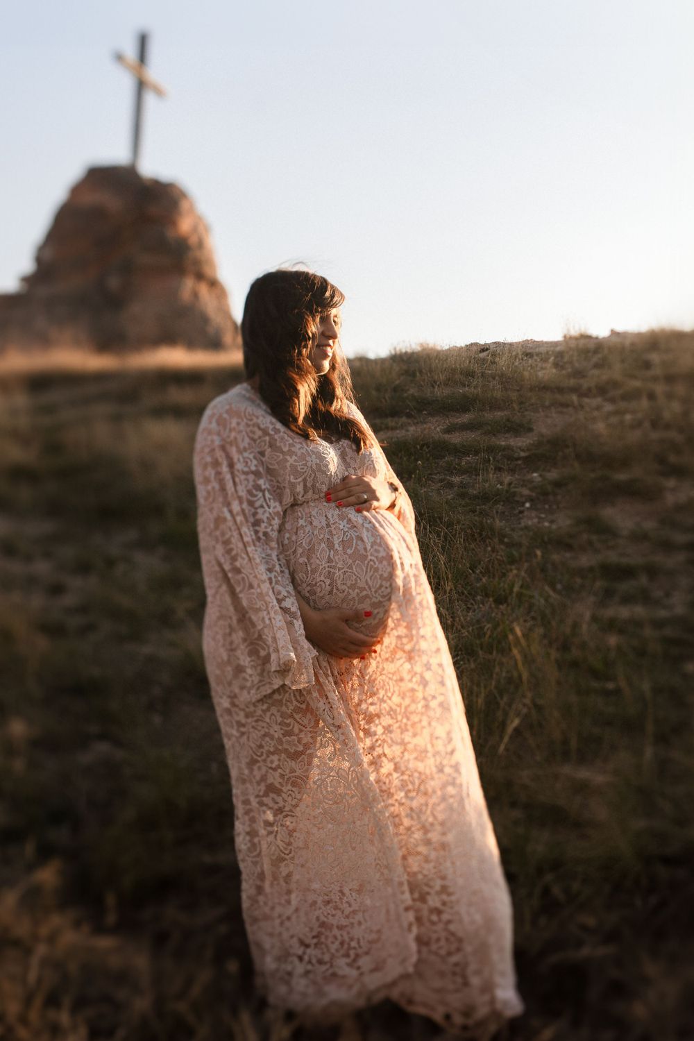 Ethereal maternity portrait with lace dress against sunset sky with cross silhouette.