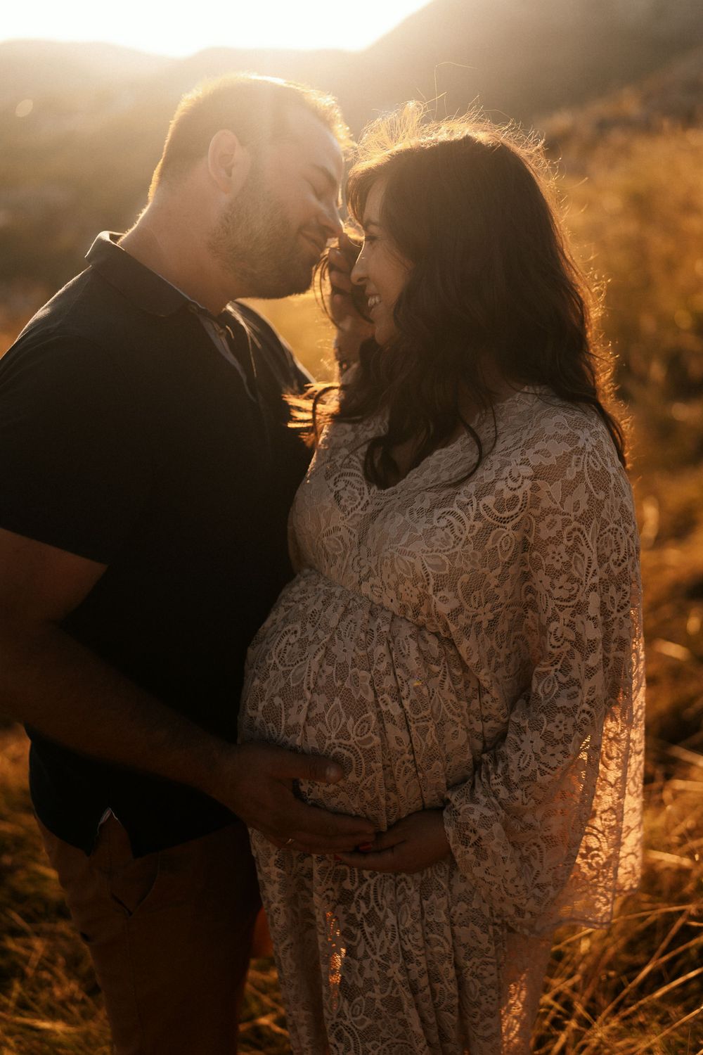 A couple embraces during a golden sunset maternity photoshoot in a field with warm backlighting.