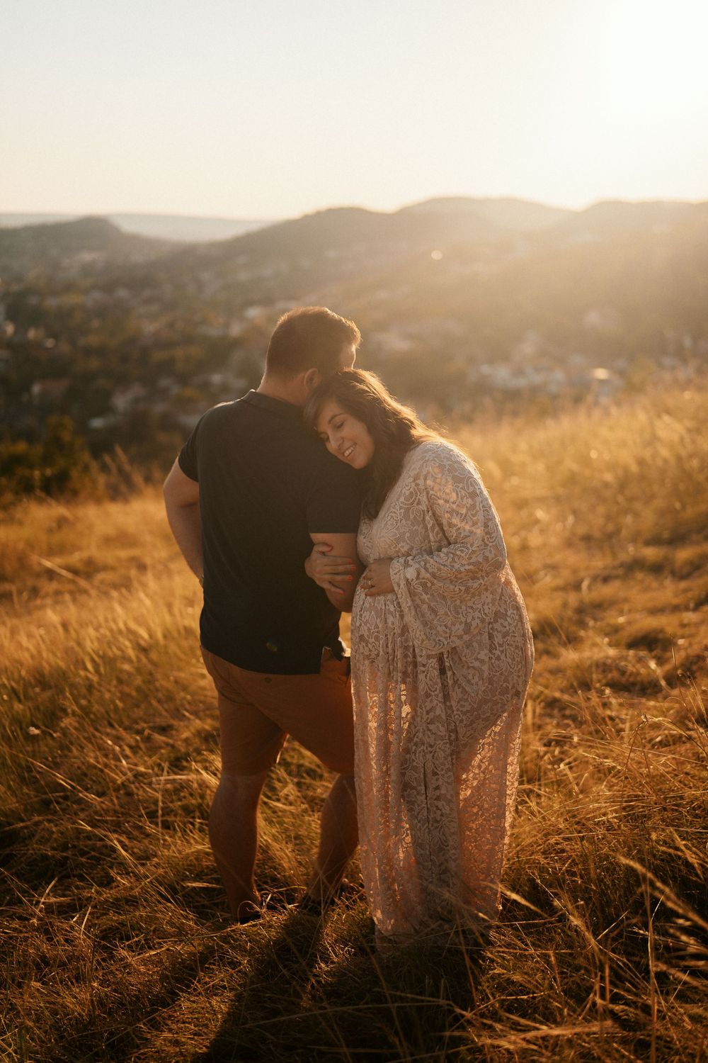 Intimate maternity moment captured on a hilltop overlook during sunset with city views in background.