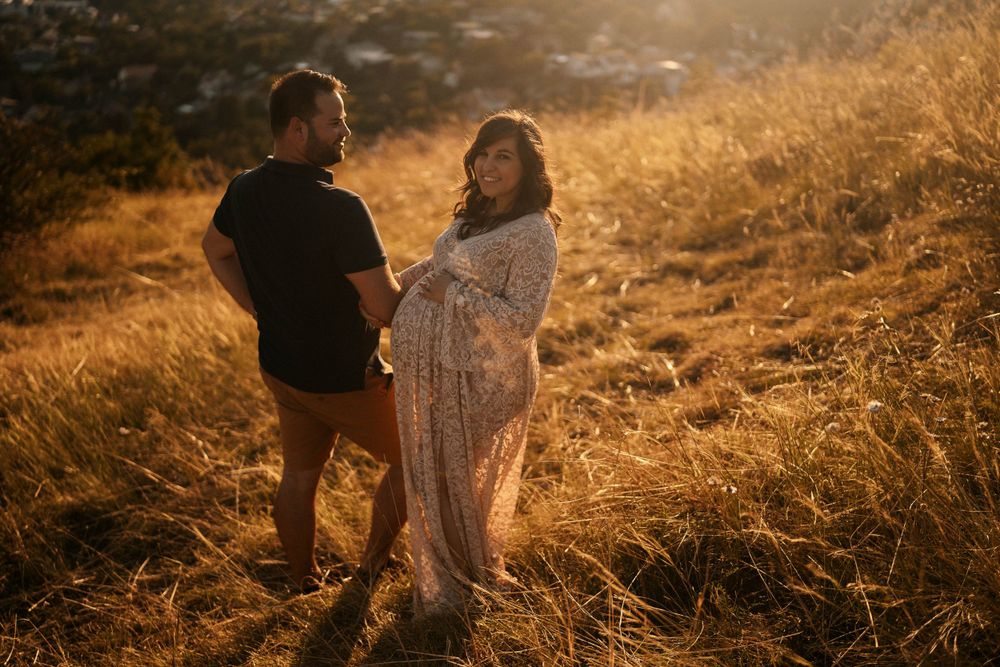 Romantic maternity photo of a couple in a lace dress and casual wear standing in golden sunlit grass.