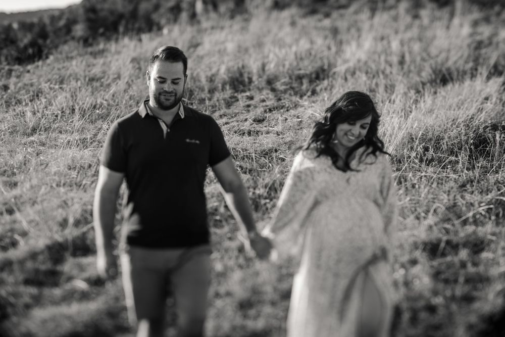 Black and white maternity photo of couple walking hand in hand on grassy hillside.