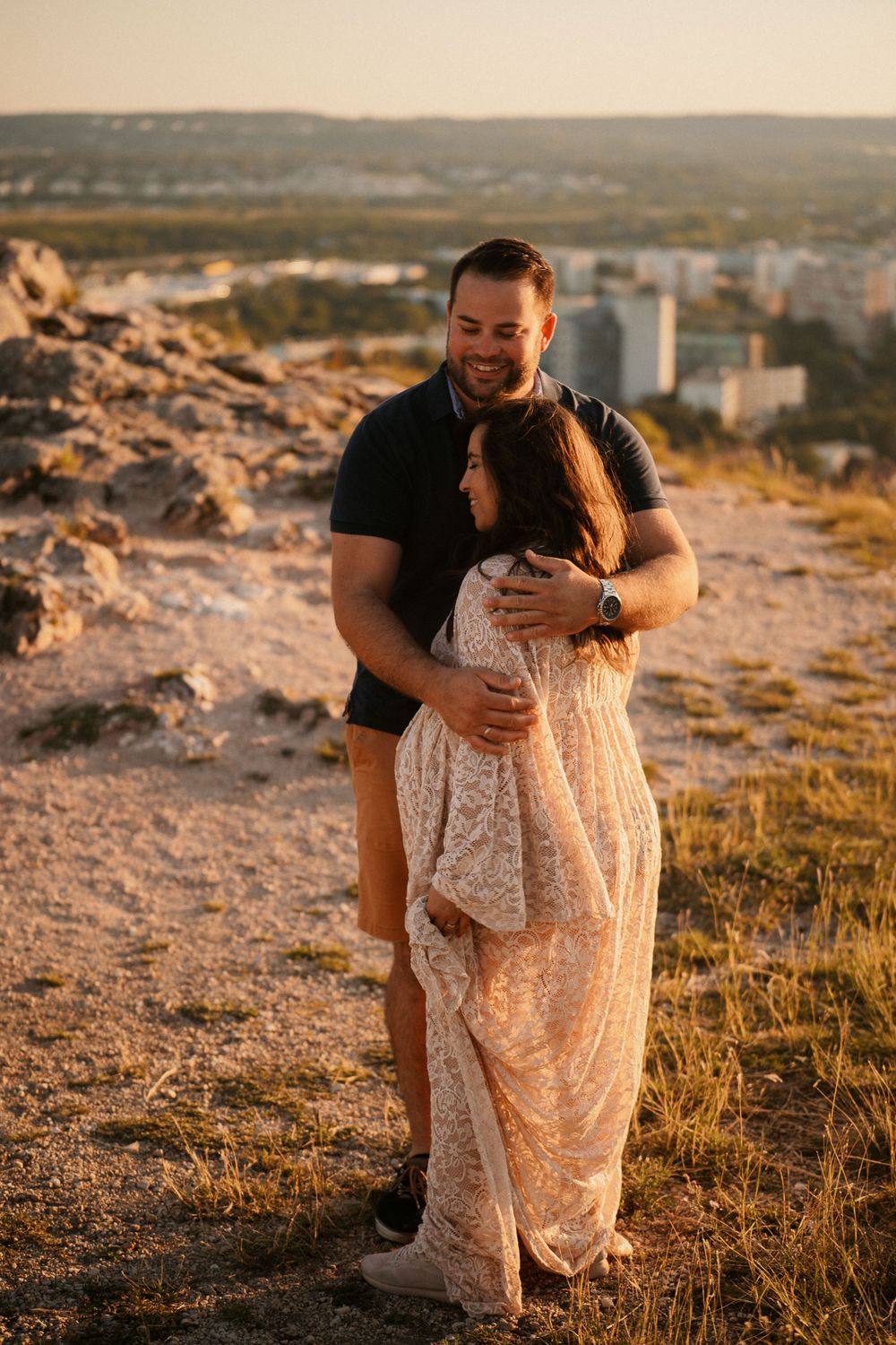 Expectant couple shares tender moment on rocky overlook with cityscape visible in background at sunset.
