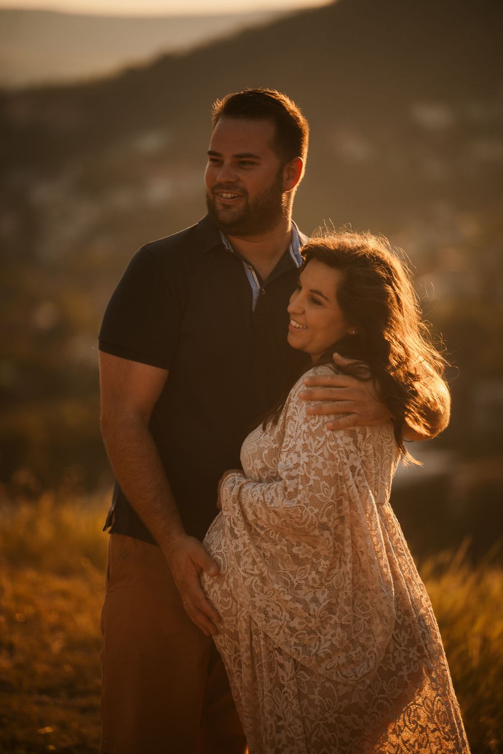 Sunset maternity portrait of couple in casual attire against mountain backdrop with golden light.