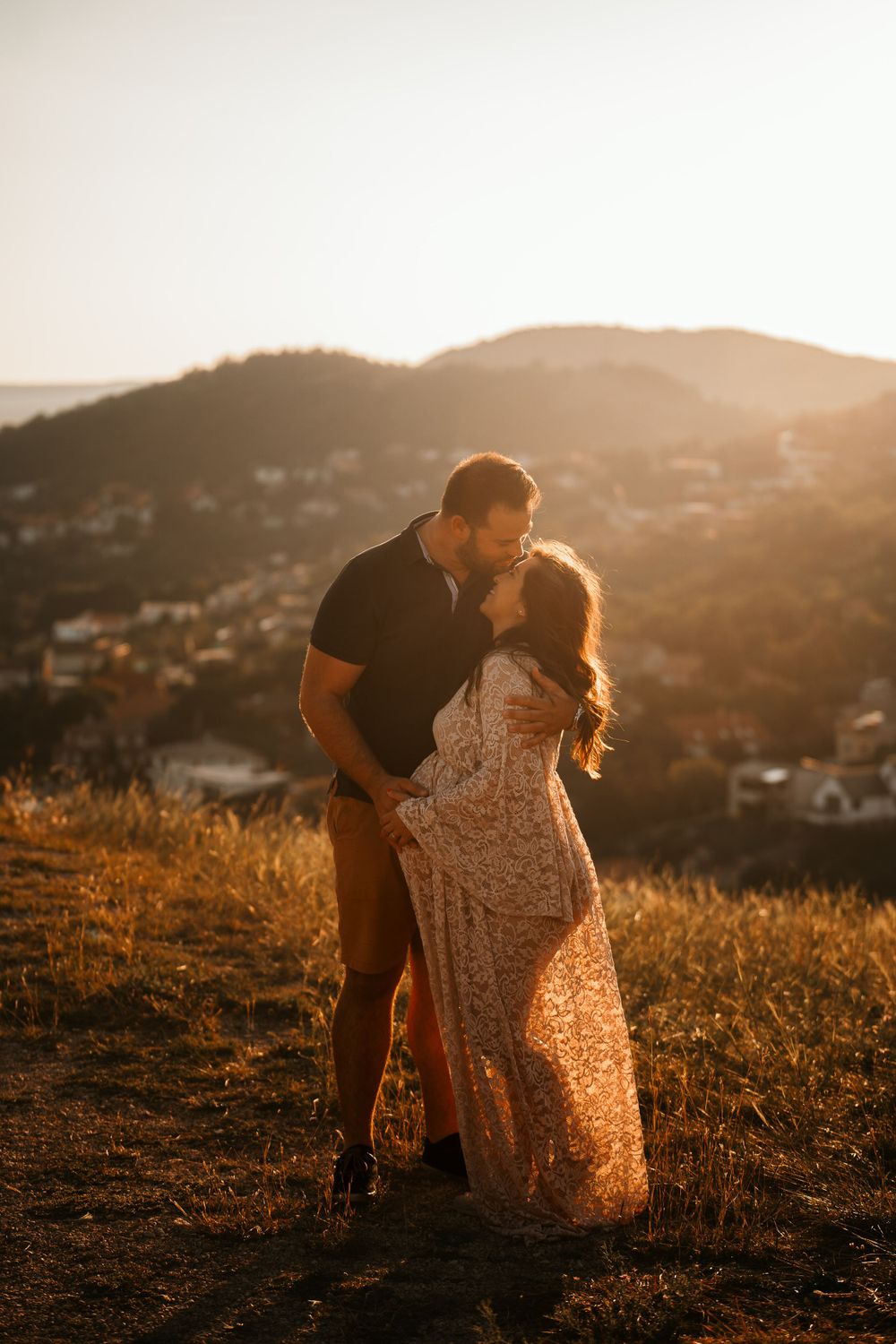 Romantic sunset maternity photo with couple on hilltop overlooking suburban landscape.
