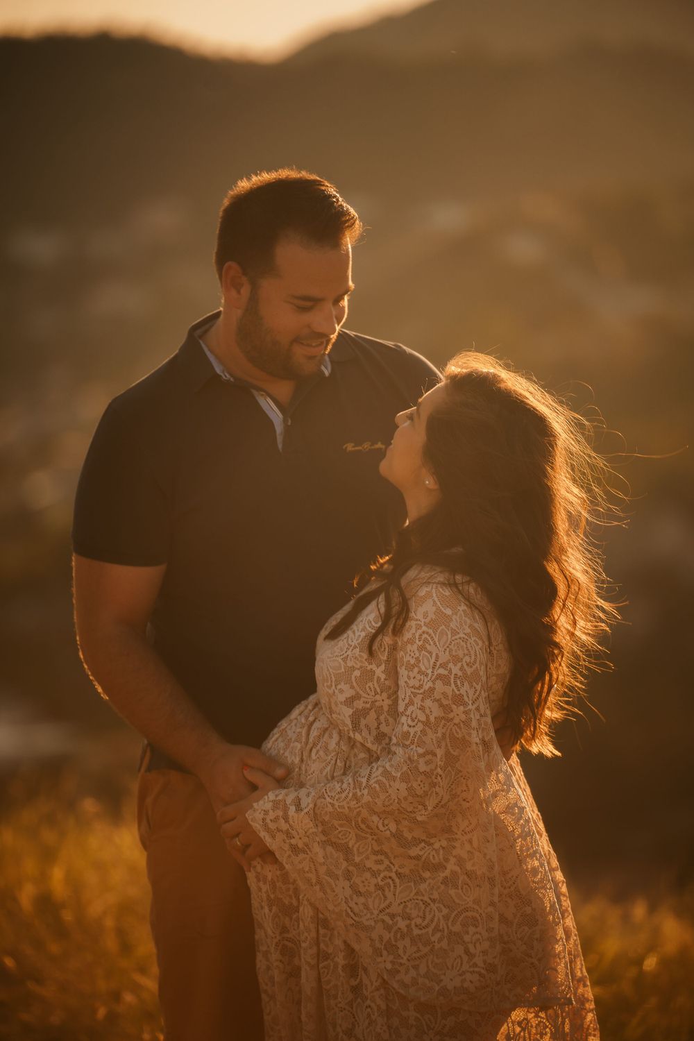 Sunset backlit maternity portrait with couple in casual attire against mountain backdrop.