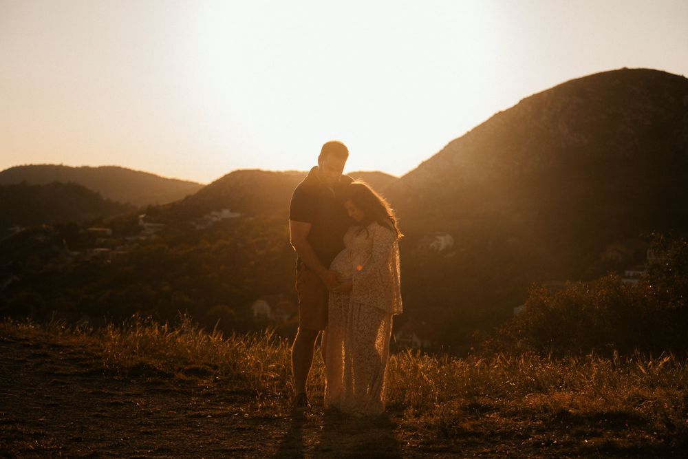 A couple stands silhouetted against a sunset over mountain peaks during a romantic outdoor photography session.