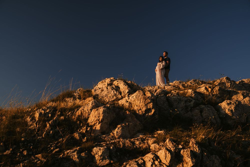A couple stands atop rocky terrain against a deep blue sky during a mountaintop photo session.