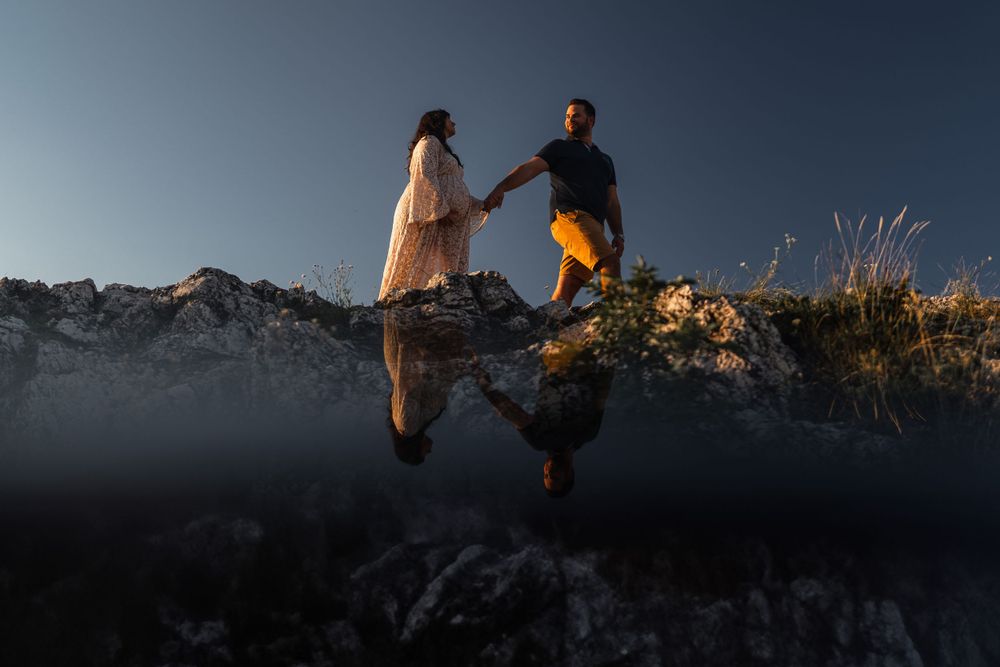 A couple's reflection appears in still water as they stand on rocky terrain at dusk.