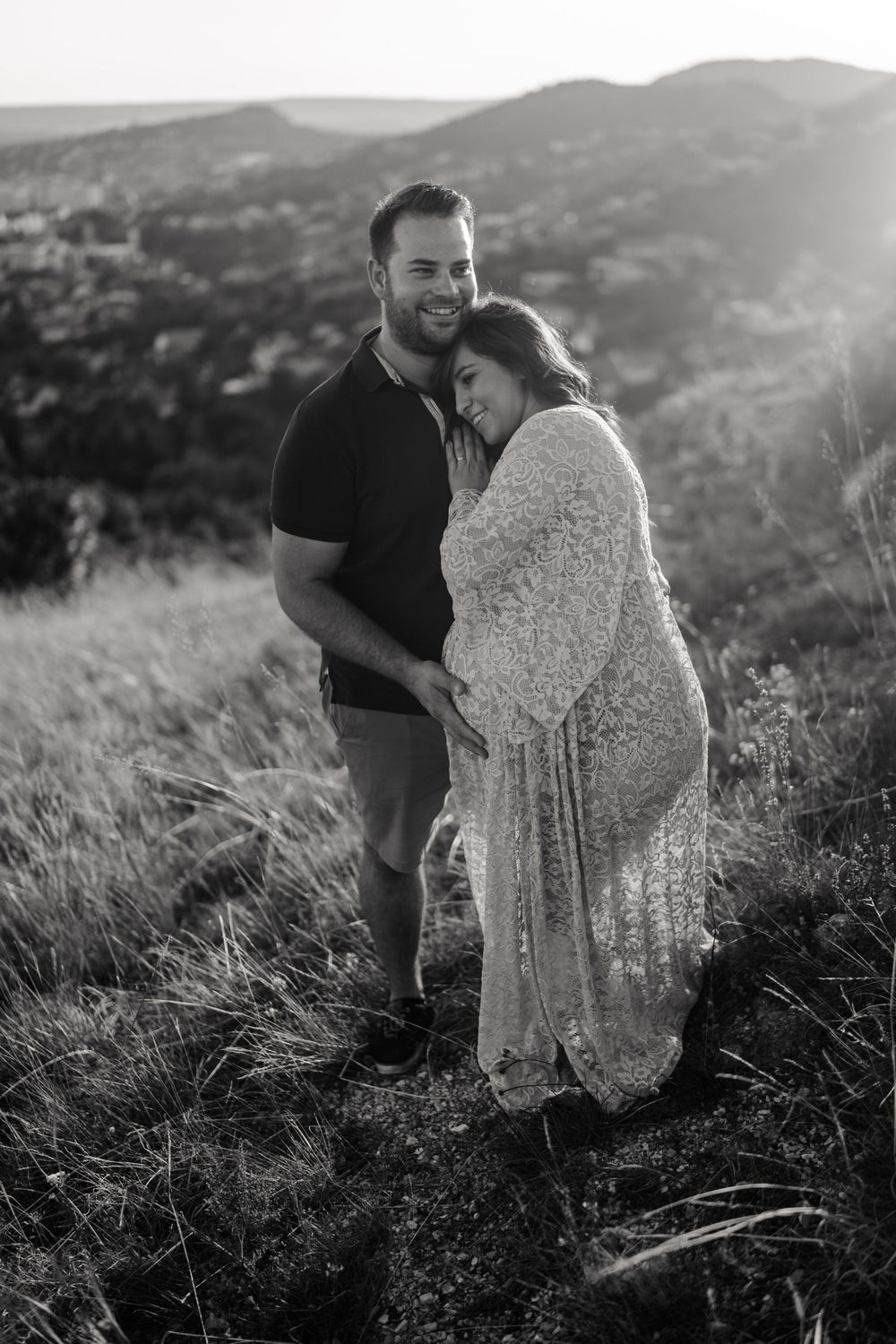 A couple embraces on a hillside overlooking a valley during a black and white maternity photo session.
