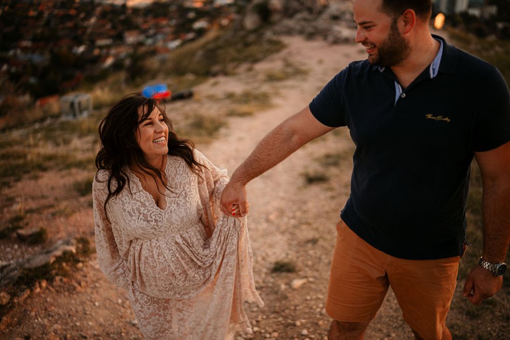A couple shares a joyful moment on a rocky hillside at sunset, with her wearing a flowing lace dress.