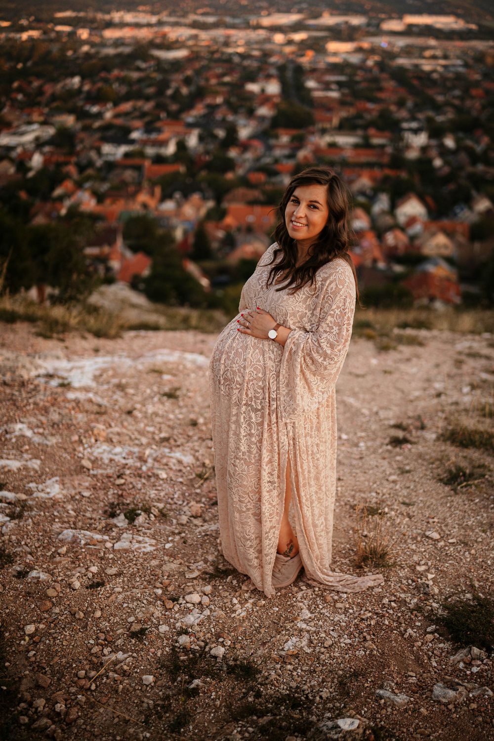 Expectant mother in a lace gown stands on a hilltop overlooking a suburban landscape at sunset.