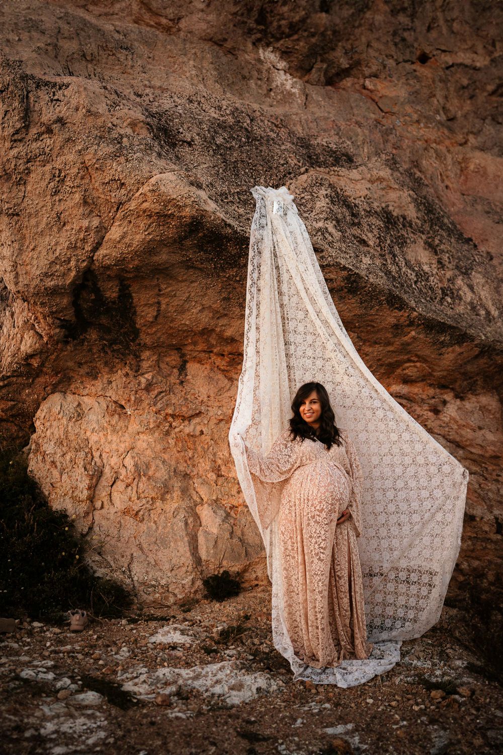 Dramatic maternity photo with white lace fabric flowing against rustic cave walls.