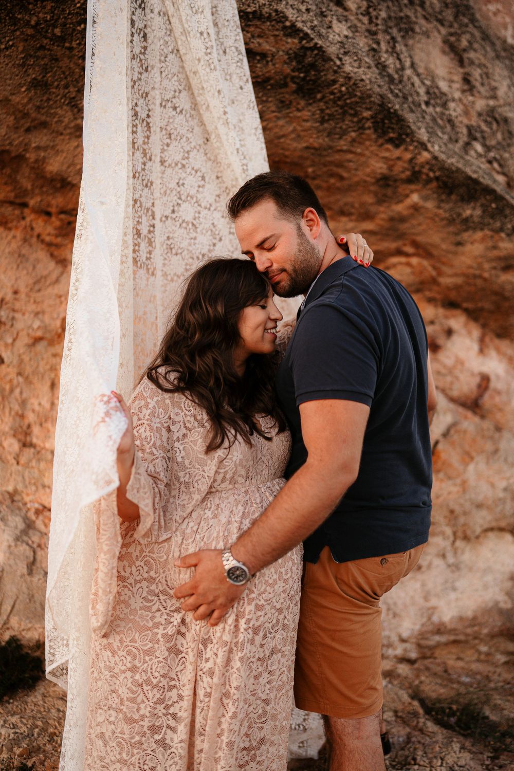 Intimate moment captured between couple against textured cave wall with flowing white lace.