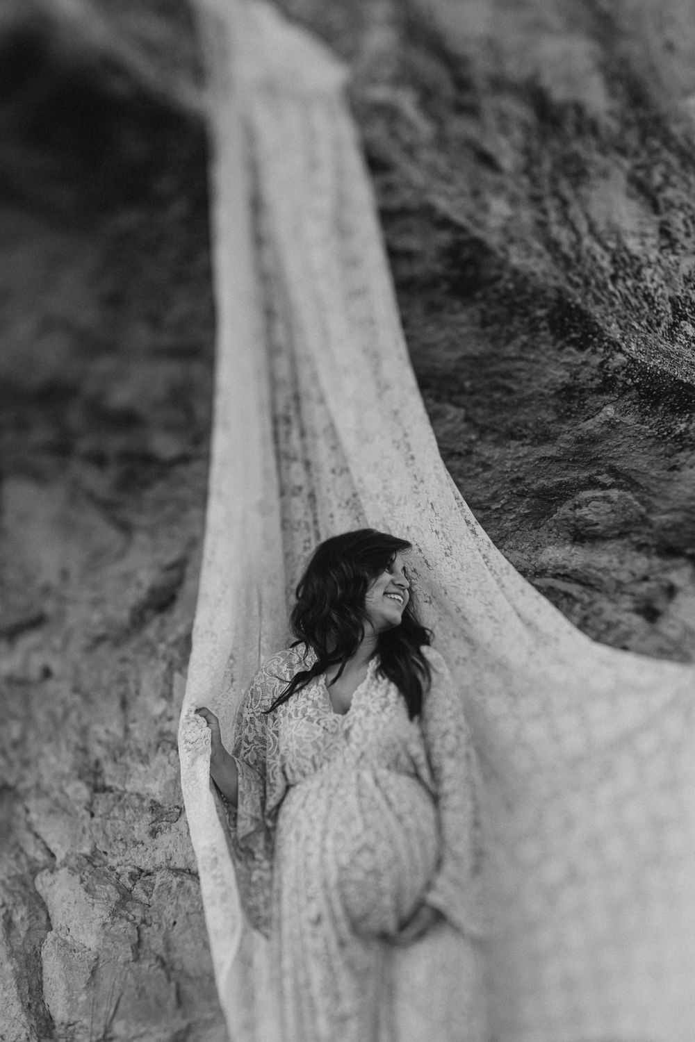 Candid black and white maternity photo featuring flowing lace fabric against rocky cave walls.