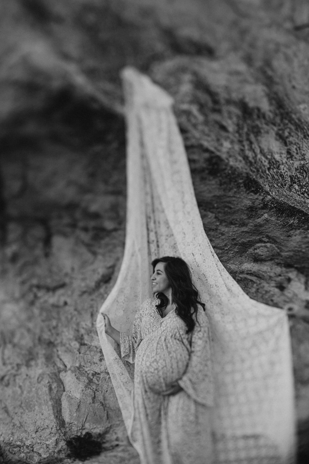 Black and white maternity portrait with flowing lace fabric in a cave setting.