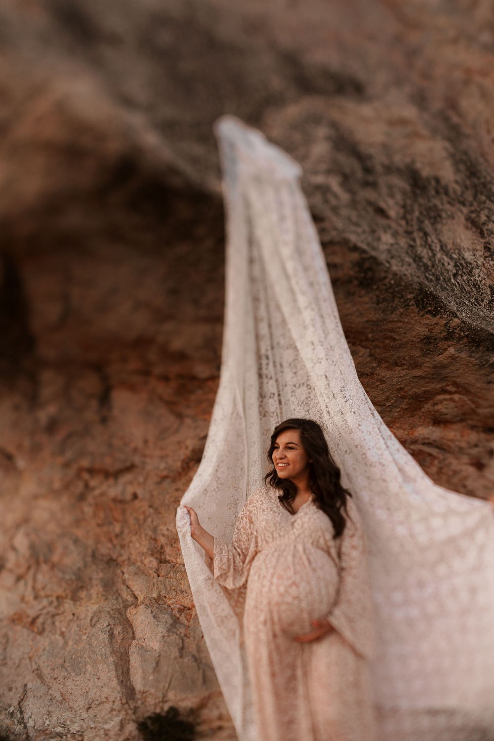 Warm-toned maternity portrait with elegant lace dress framed by natural cave walls.