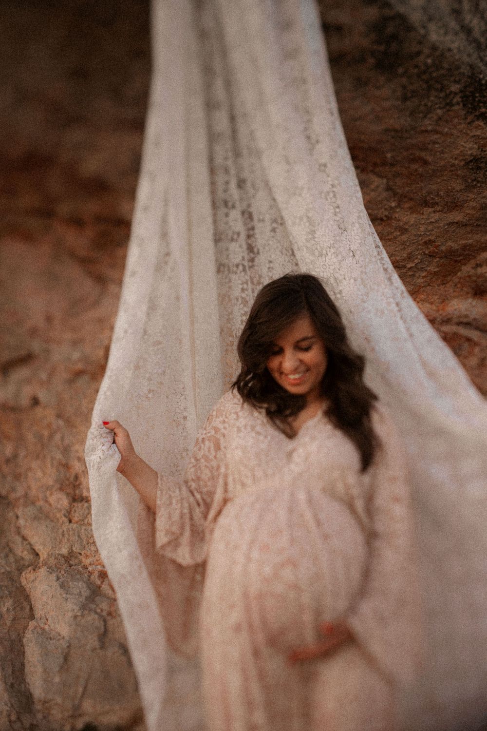 A dreamy maternity portrait showing a pregnant woman in a white lace dress against flowing fabric in a soft, moody setting.