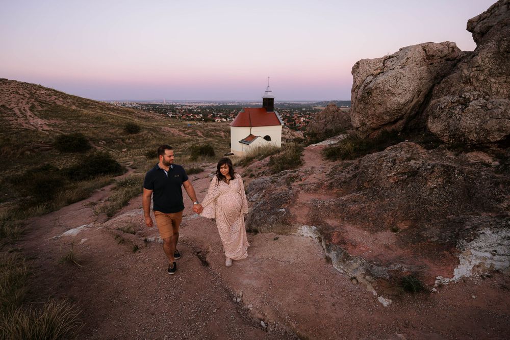 A couple walks along a rocky path at sunset with a white church visible in the distance.