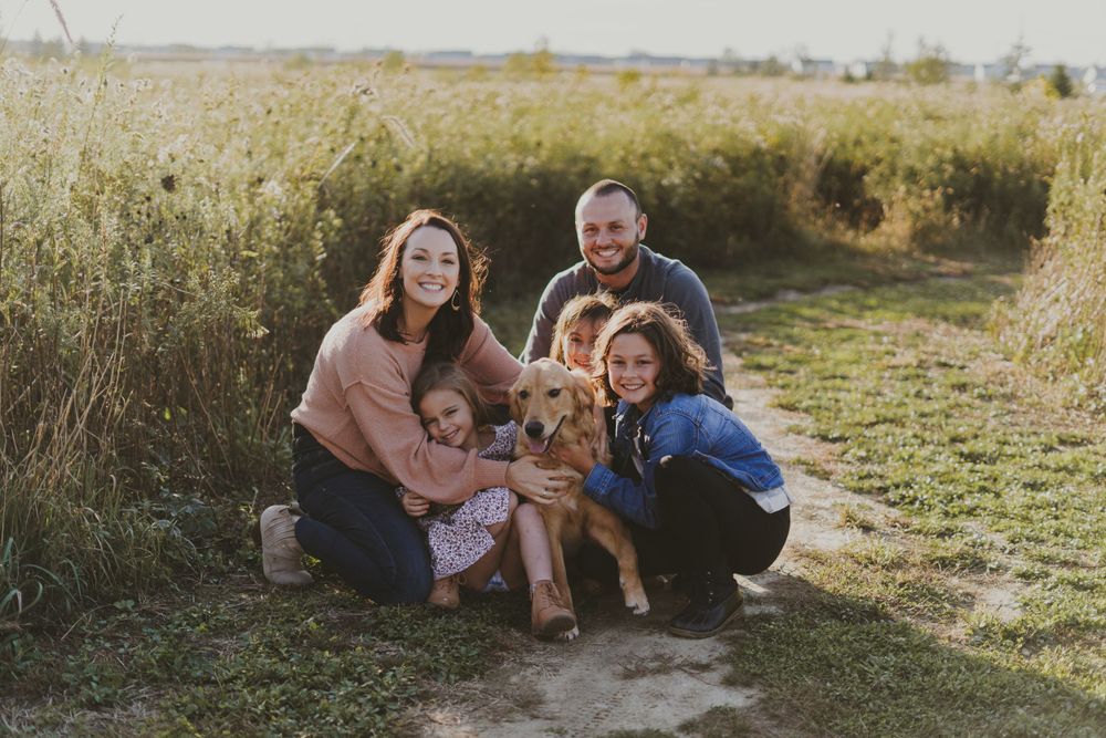 A family and their dog pose together for a casual portrait in a golden field at sunset.