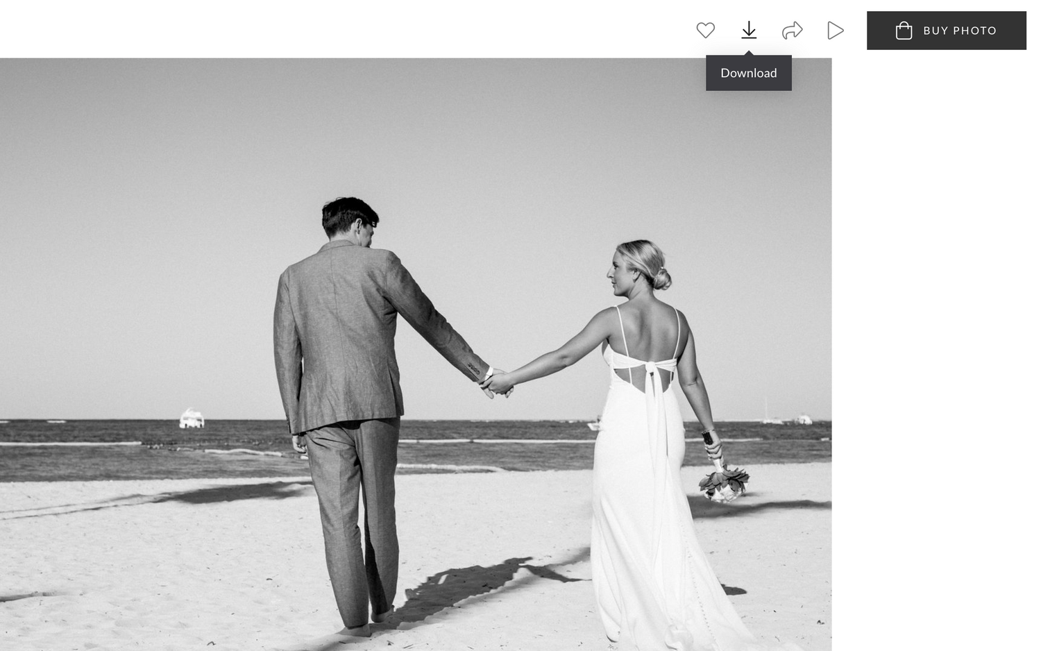 Black and white photo of newlywed couple holding hands while walking along a sunny beach shoreline.