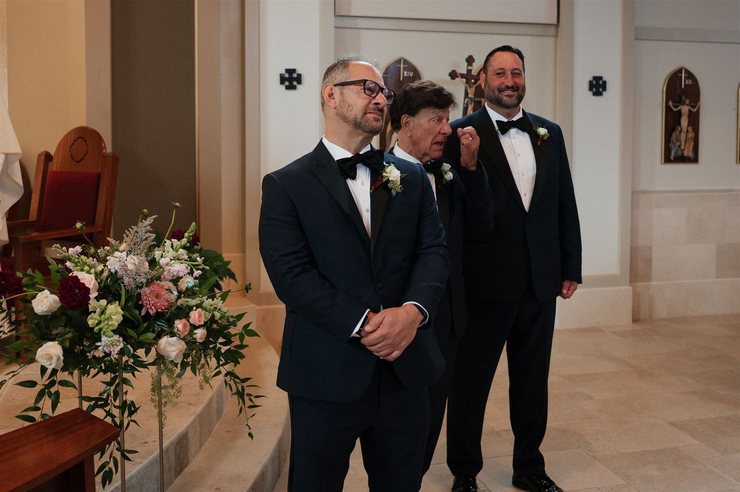 Groomsmen in black tuxedos standing at altar with floral arrangements.