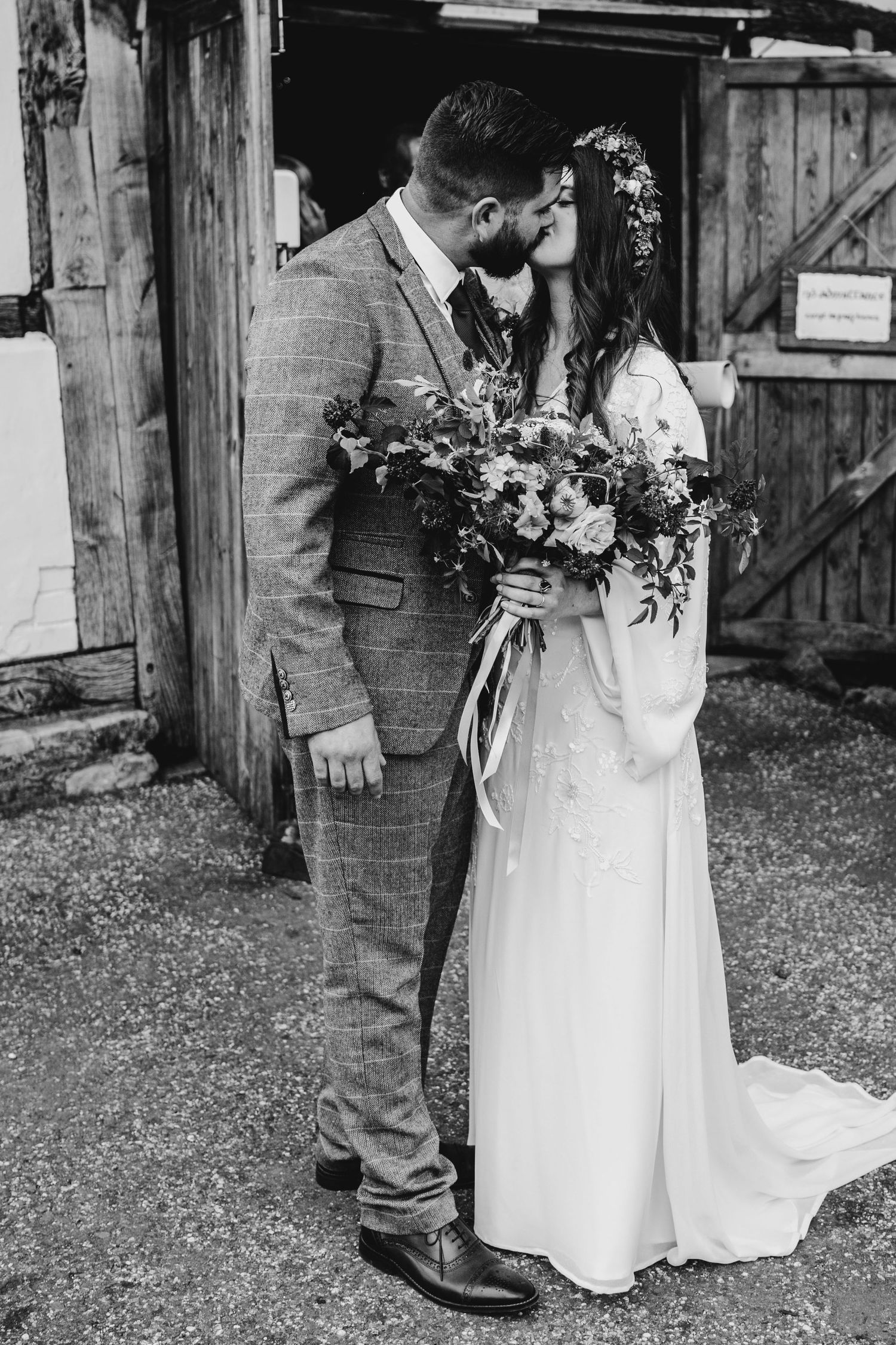 Black and white portrait of a couple in wedding attire standing by a rustic barn.