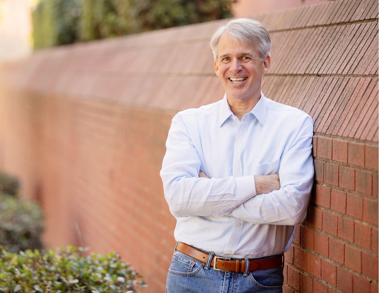 Smiling middle-aged man leaning against a brick wall with arms crossed in outdoor setting.