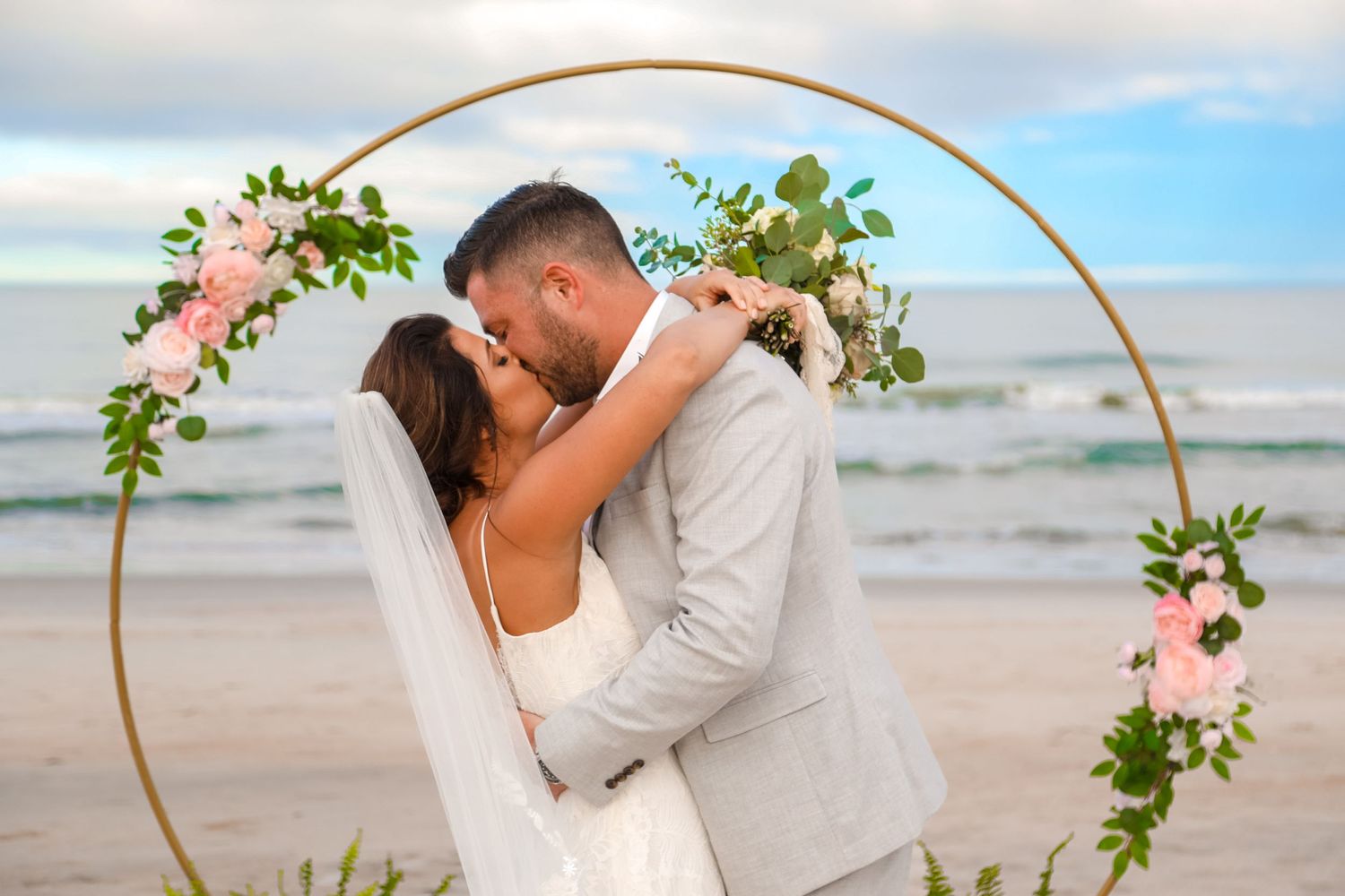 Beach wedding ceremony with floral arch and ocean backdrop.