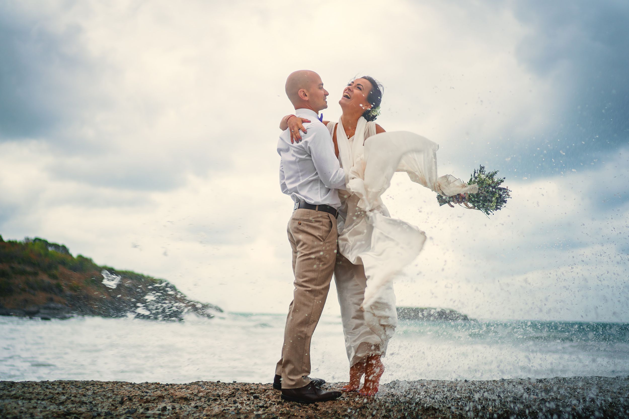 A joyful seaside moment captured with flowing white dress and bouquet against ocean waves.