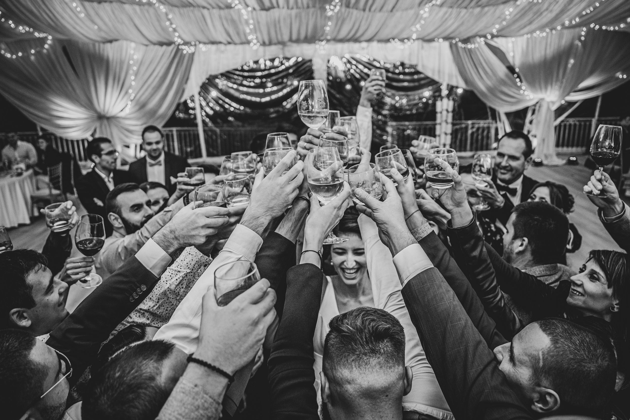 A black and white photo captures a celebratory toast with many raised glasses at a wedding reception.