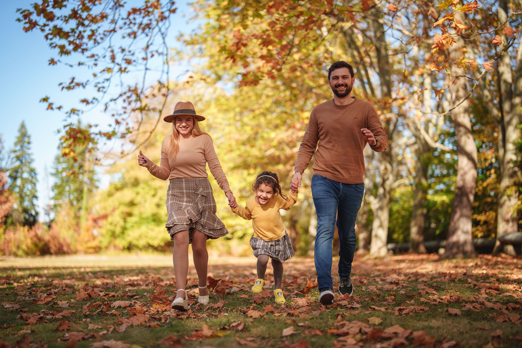 A happy family walks together through fallen autumn leaves in a sunlit park with golden foliage.