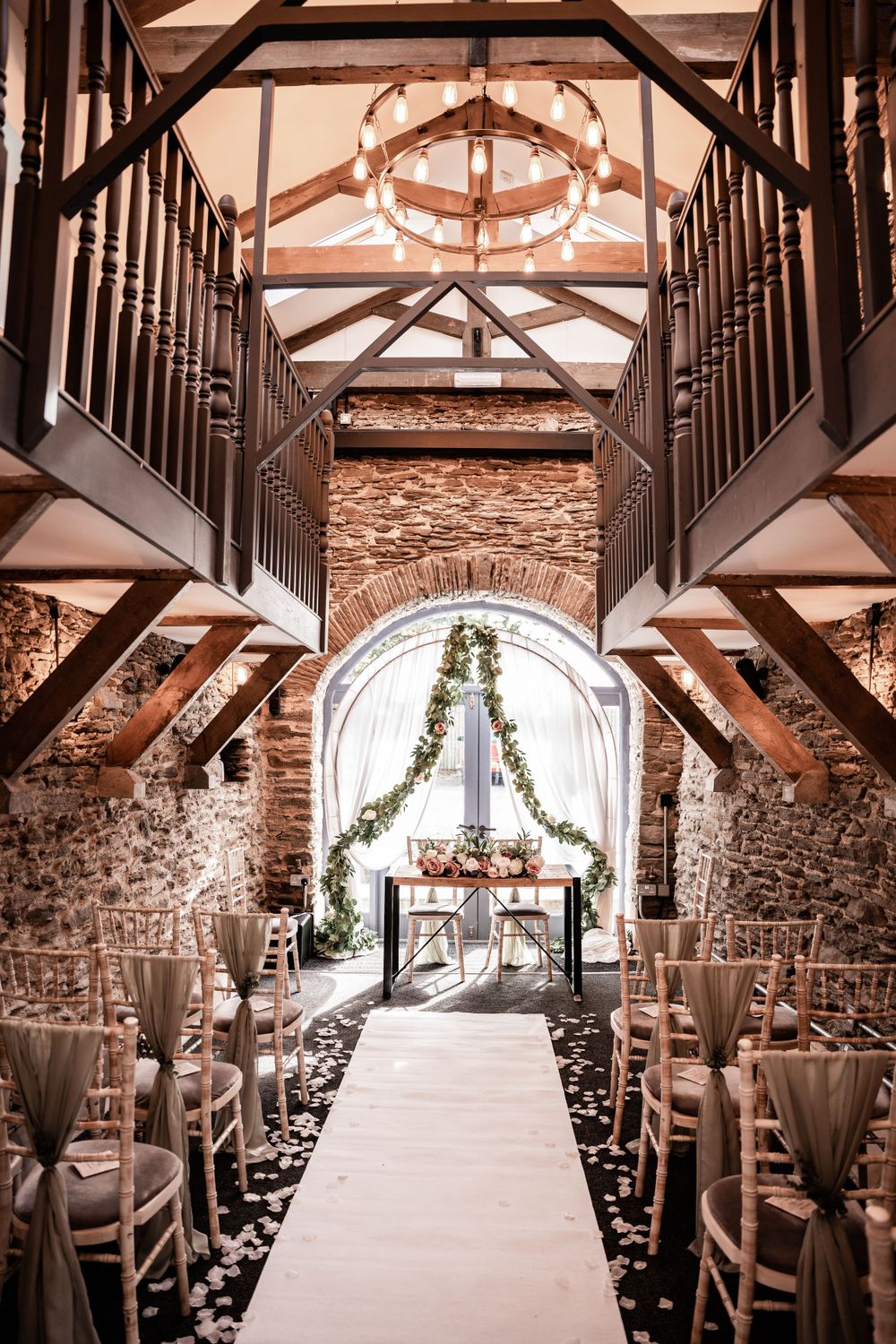 Wedding ceremony aisle in rustic barn with wooden beams and hanging chandelier.