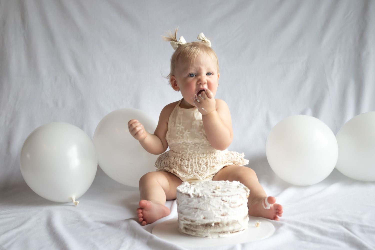 Baby girl eating cake on her first birthday in a white dress with white balloons and a white backdrop