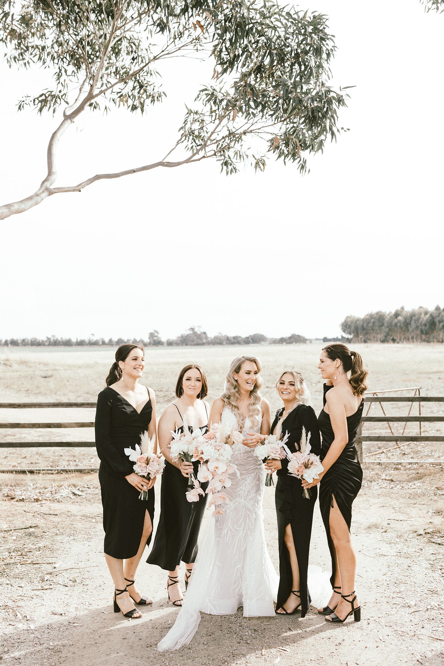 Bridal party in elegant black dresses pose with bouquets under eucalyptus tree.