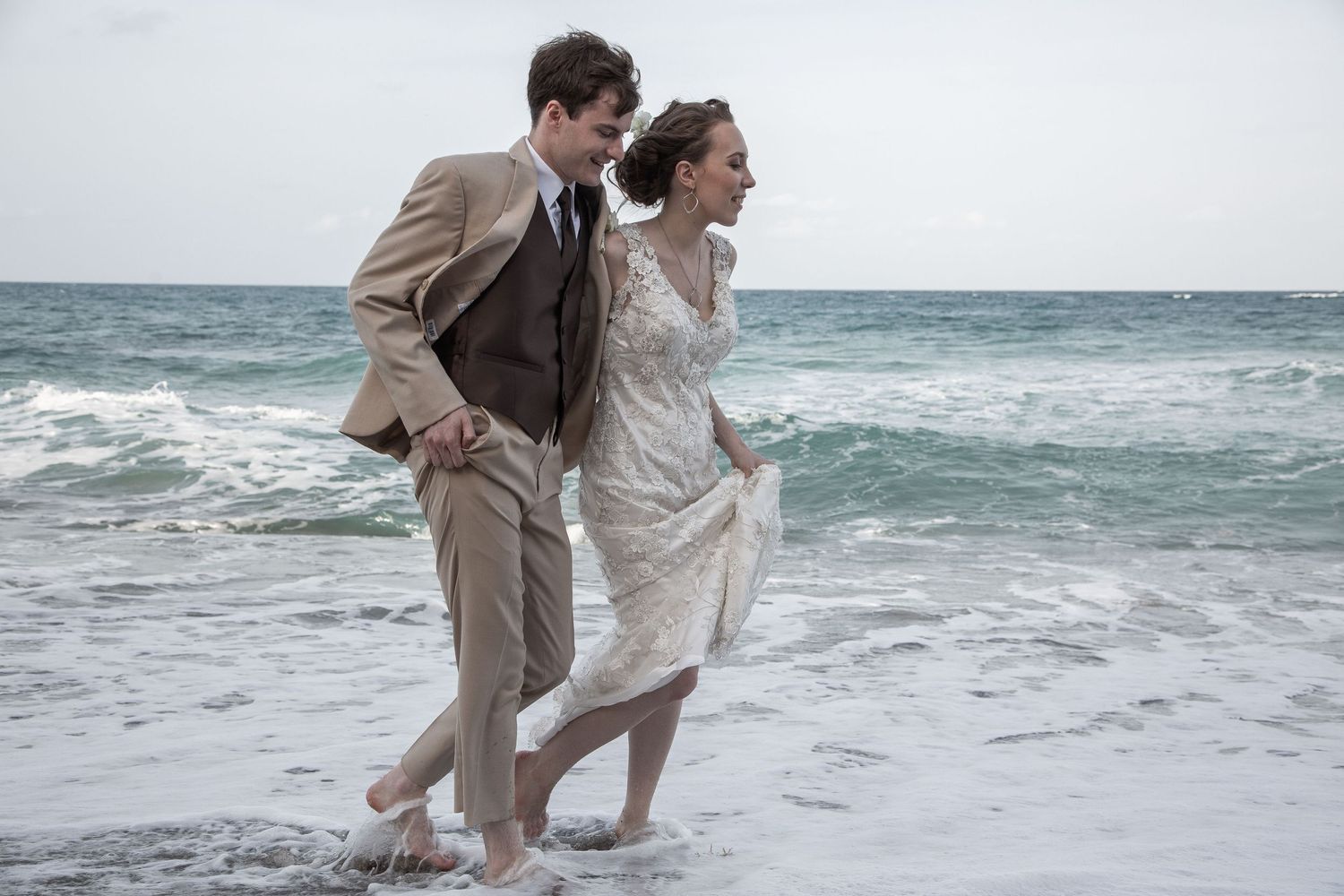 A couple walks along a beach shore in formal attire as waves crash behind them.